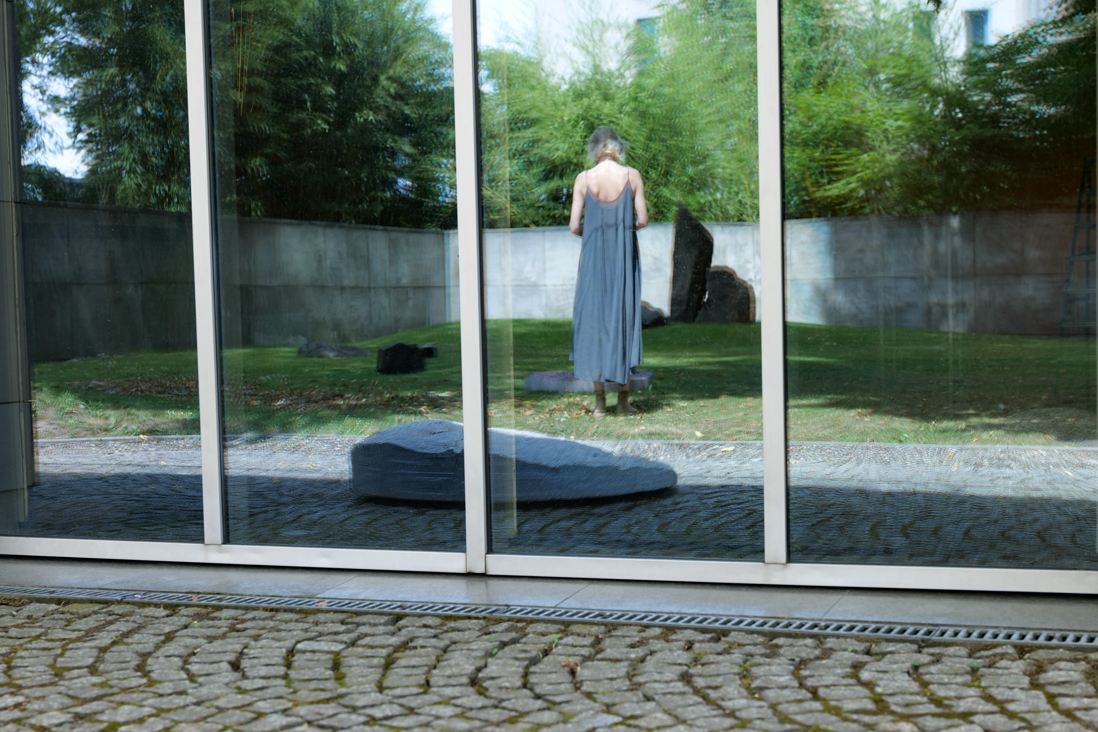Window reflection at the Manggha Museum showing Ewa Wes Wesołowska’s sculptures in the Japanese garden and a woman standing in the garden, seen from the back.